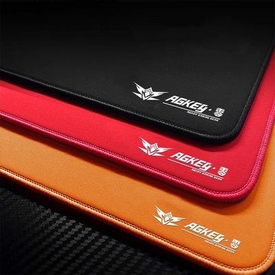 Red AGKey Zero gaming mouse pad, stacked with black and orange pads, featuring a white logo and stitched edges.